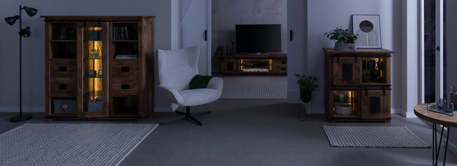 RAILWAY-woonruimte ’s nachts met verlichte vitrinekasten, een tv-lowboard en een gezellige fauteuil in industriële stijl.