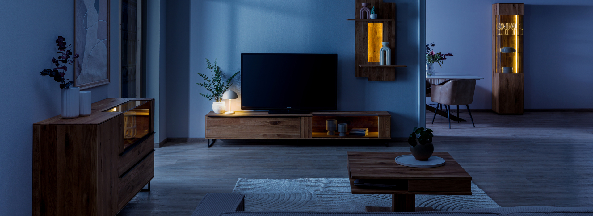 Woonkamer ’s nachts met wilde eik, een warm verlichte vitrinekast, wandplanken en een tv-lowboard in een rustige sfeer.