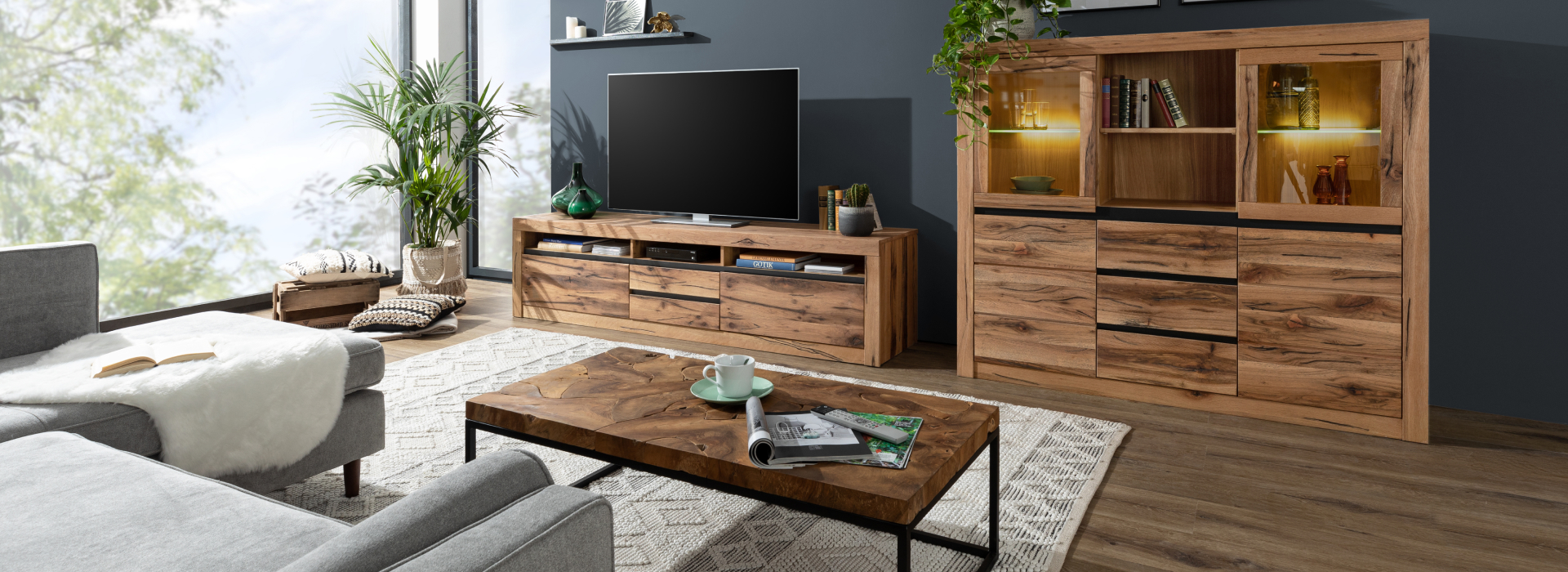 Moderne woonkamer met massief houten meubels, verlichte vitrinekast, tv-lowboard en salontafel met metalen onderstel.