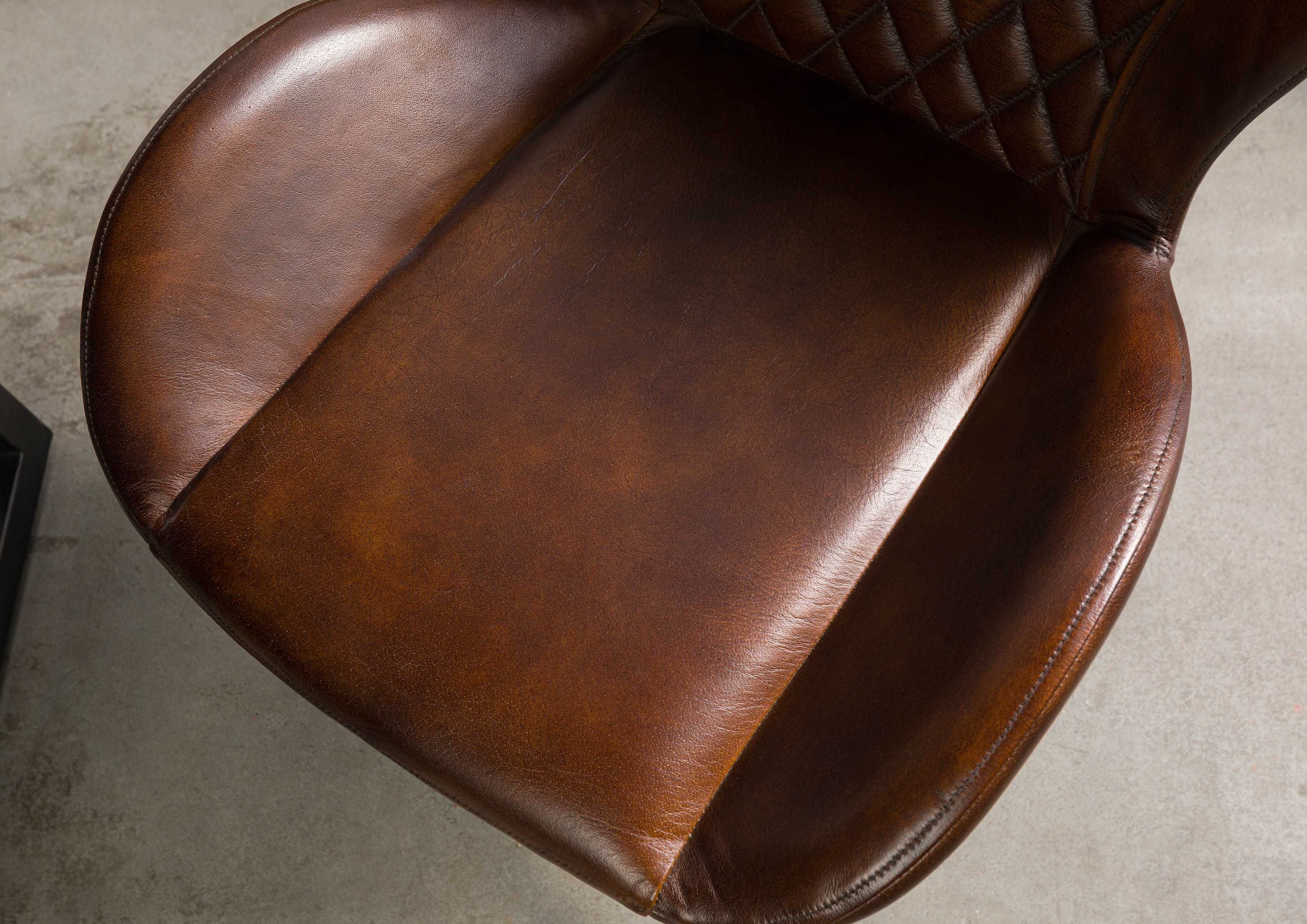 Detailfoto van een bruine leren stoel met een zachte zitting, armleuningen en  doorstikwerk op de rugleuning.