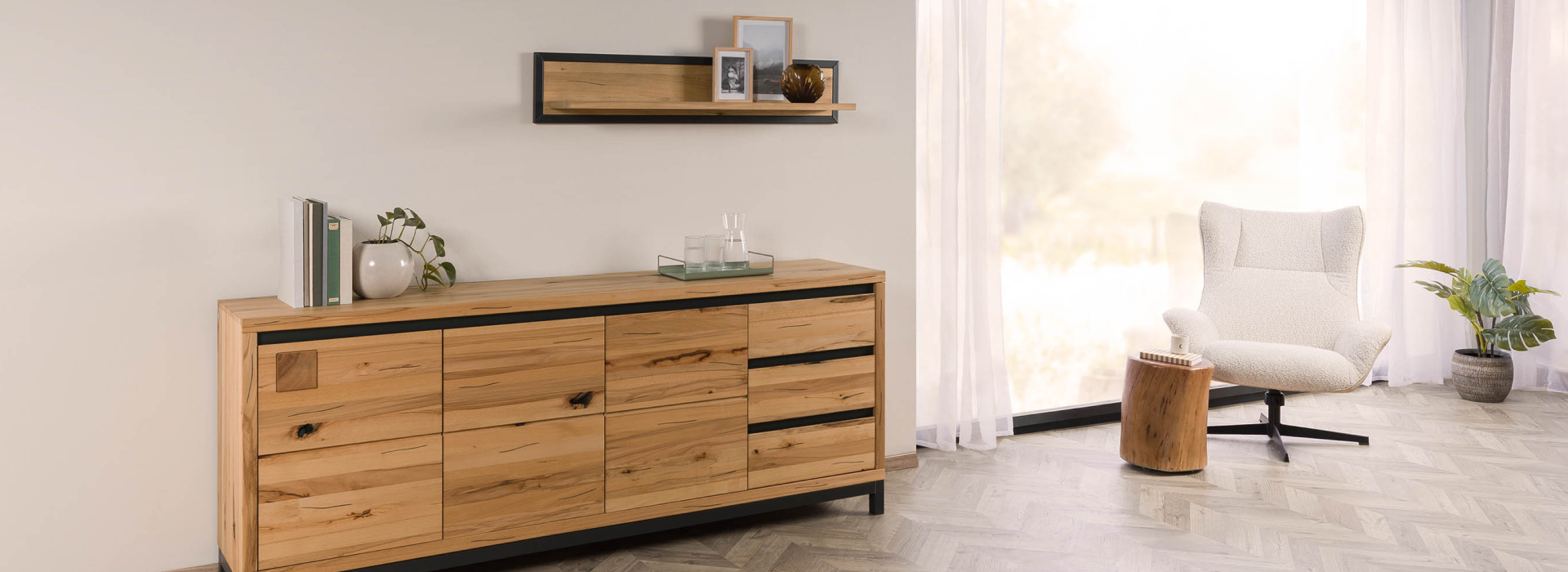Lichte woonruimte met een sideboard van kernbeuk, zwarte accenten, een wandplank en een gezellige fauteuil bij het raam.