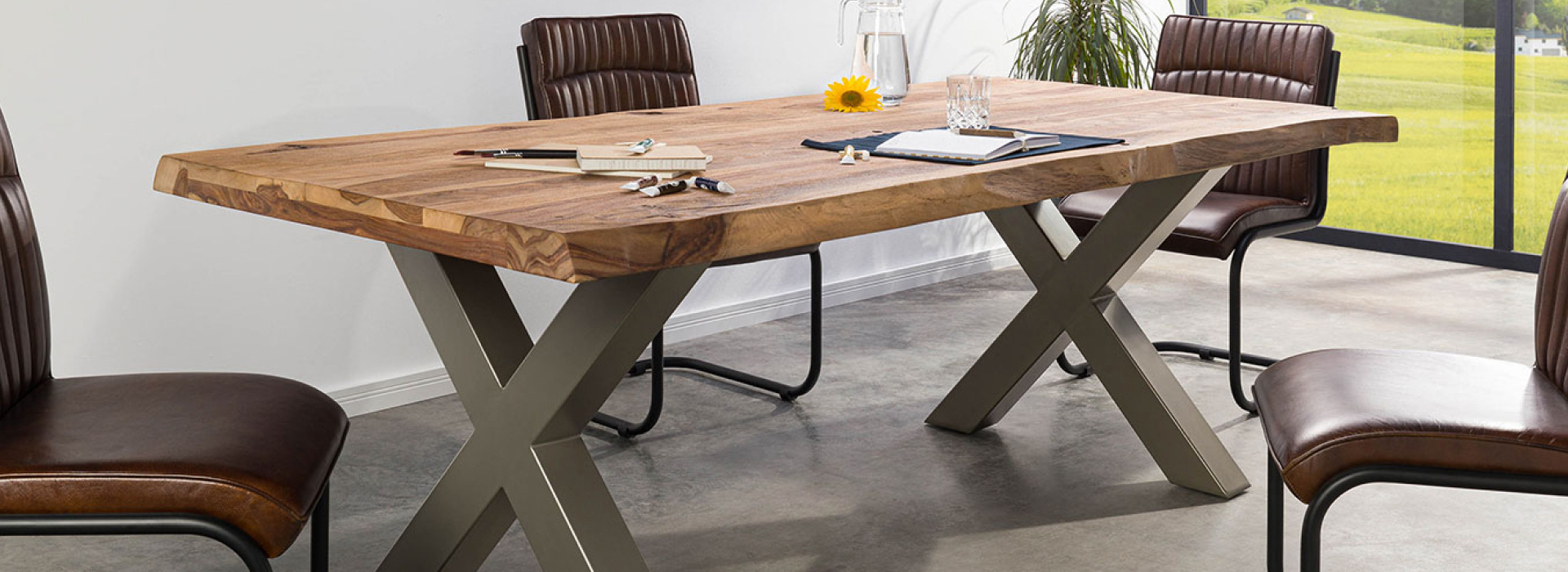 Boomtafel met massief houten blad en twee X-vormige poten in mat zilver, omringd door bruine stoelen.