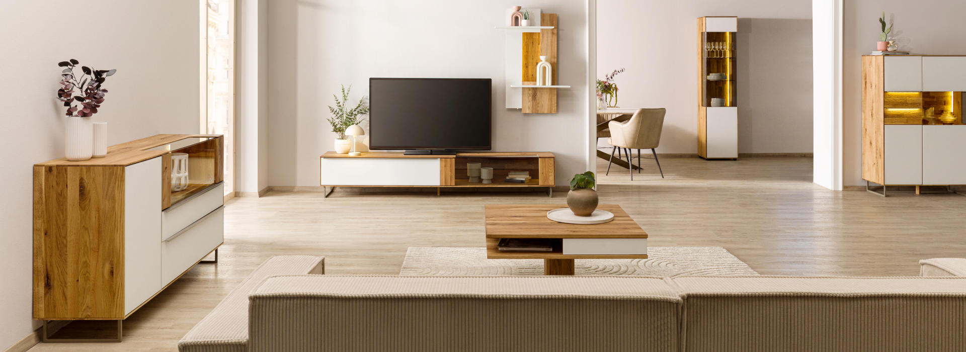 Lichte woonkamer met meubels van wild eiken, een tv-meubel, een dressoir en een warm verlichte wandplank als blikvanger.