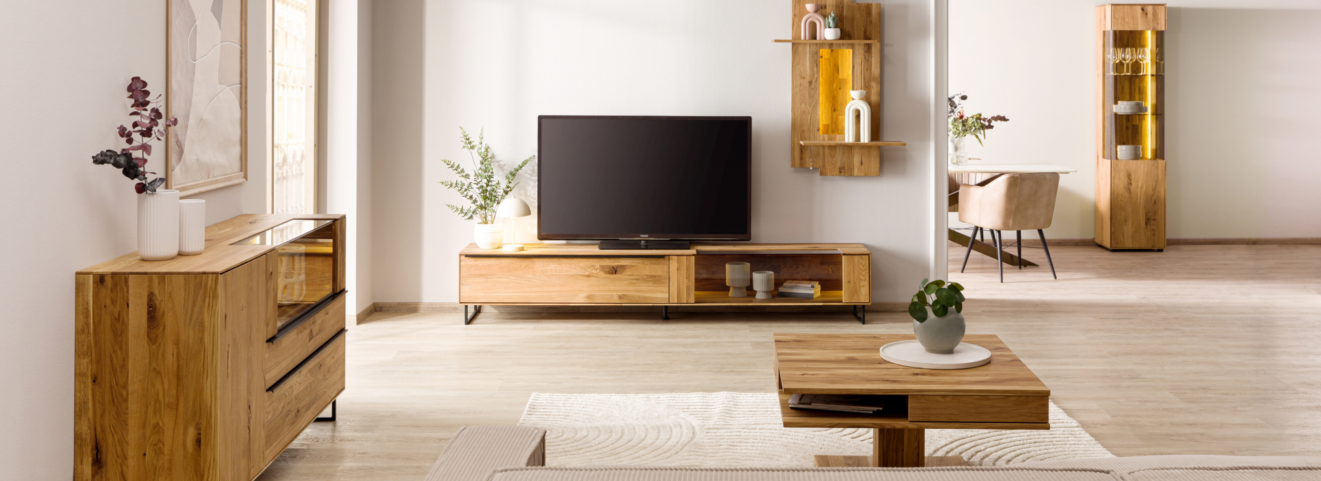Lichte woonkamer met meubels van wild eiken, een tv-meubel, een dressoir en een warm verlichte wandplank als blikvanger.