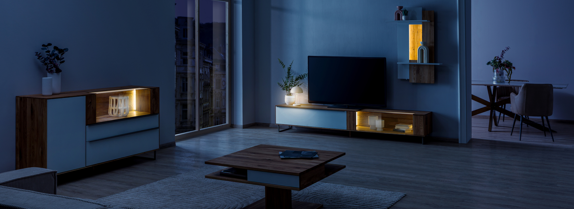 Woonkamer ’s nachts met wilde eik, een warm verlichte vitrinekast, wandplanken en een tv-lowboard in een rustige sfeer.